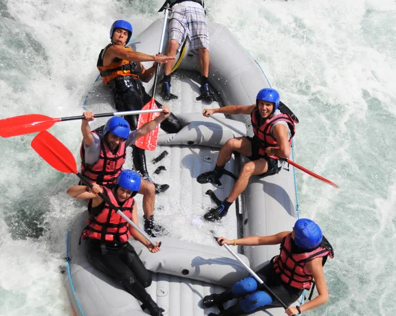 Six people in a raft paddling through whitewater rapids, wearing helmets and life jackets.