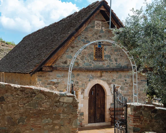 Stone chapel with arched doorway, thatched roof, and trees under a partly cloudy sky.