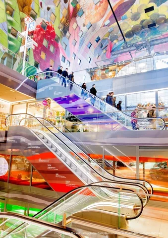 People on colorful escalators inside a modern, brightly decorated shopping mall.