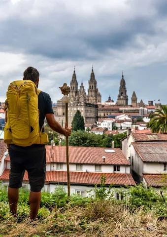 Hiker with yellow backpack overlooking city with cathedral under cloudy sky.