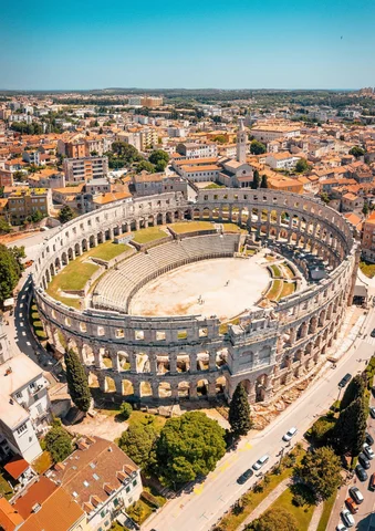 Ancient Roman amphitheater surrounded by city buildings under a clear blue sky.