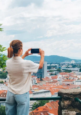 Woman taking a photo of a cityscape with red rooftops and distant hills.