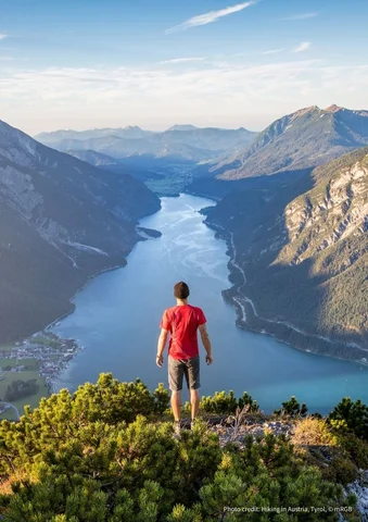Person in red shirt standing on a mountain overlooking a river valley and distant peaks.