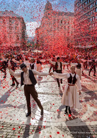 Dancers in traditional costumes performing in a city square with red confetti falling.