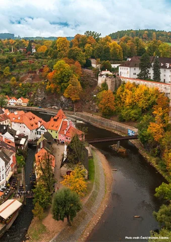 A river curves through a town with red-roofed buildings and colorful autumn trees.