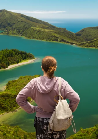 Woman with a backpack overlooks a turquoise lake surrounded by green hills.