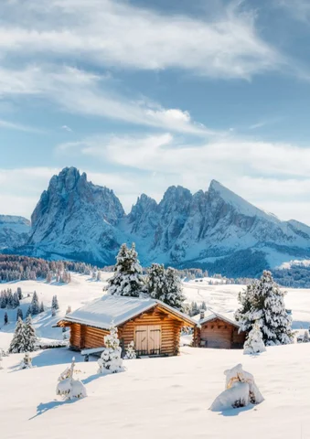 Snow-covered cabins and trees with towering mountains in the background under a blue sky.