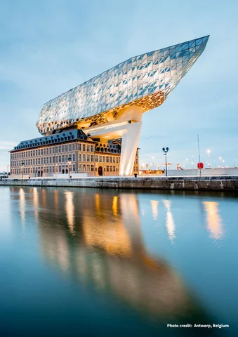 Modern glass building atop historic structure reflected in calm water at dusk.