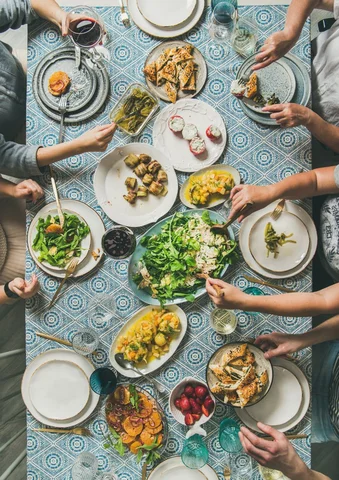 People sharing a meal with salads, vegetables, and wine on a patterned tablecloth.