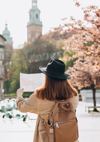 Woman with a backpack and hat holding a map, standing near blooming trees in a city.