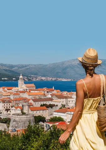 Woman in a yellow dress overlooking a coastal town with red rooftops and mountains.