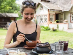 Woman enjoying traditional Seto food outdoors at Taarka Taro KöögiKono restaurant in Estonia, with rustic farm buildings in the background.
